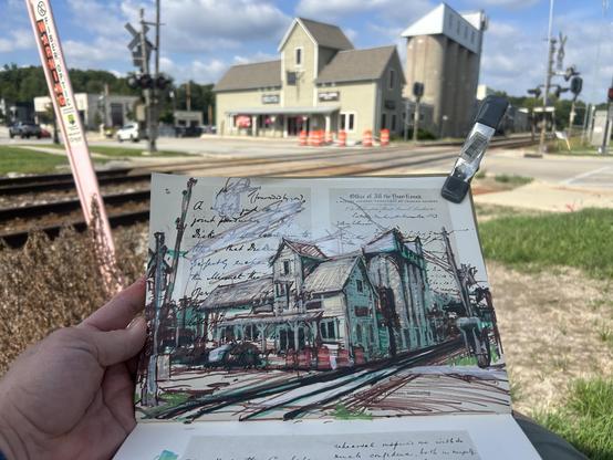 An outdoor drawing of a train crossing and an old silo that has some businesses in it. I’m sitting in a lawn chair and drawing in my salvaged book.