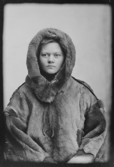 A young woman stares impassively at the camera while wearing a very thick arctic-style fur coat.